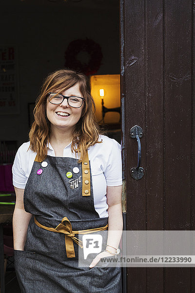 Woman with long brown hair wearing apron and glasses standing in an open doorway  smiling.