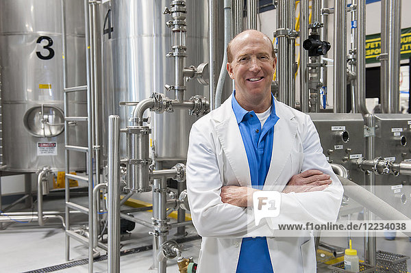 Caucasian male technician in a white coat standing near large stainless steel processing tanks in a bottling plant that makes flavoured water .
