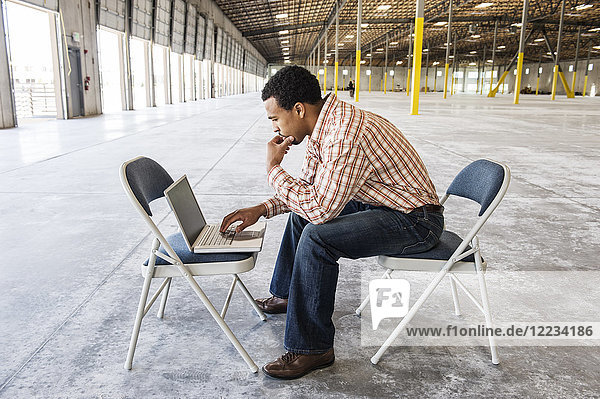 Black man working on lap top computer in front of loading dock doors in a new warehouse.