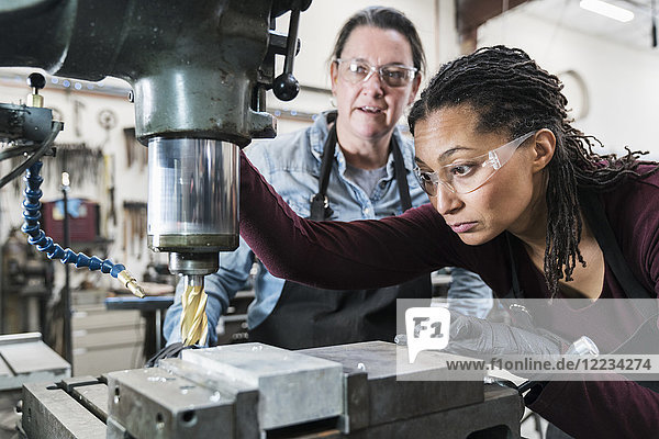 Two women wearing safety glasses standing in a metal workshop  working on metal drilling machine.
