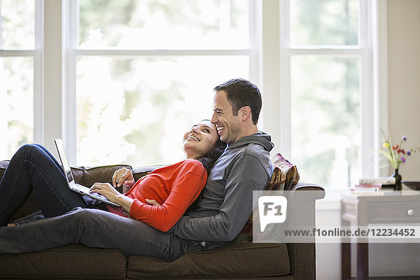 Caucasian man and mixed race Caucasian woman using a lap top computer while sitting on a couch in their home.