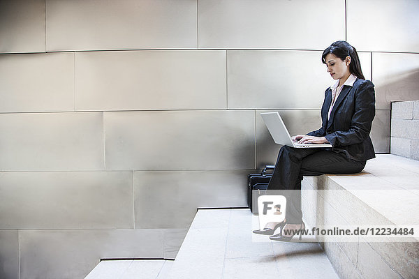Eine hispanische Geschäftsfrau sitzt auf einer Treppe in der Lobby eines großen Bürogebäudes.