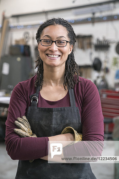 Frau mit braunen Haaren  die eine Brille und eine Schürze trägt  steht mit verschränkten Armen in der Metallwerkstatt und lächelt in die Kamera.