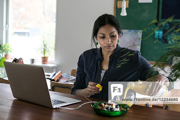Weibliche Design-Profis gießen Saft in Glas mit Laptop auf dem Tisch im Home-Office