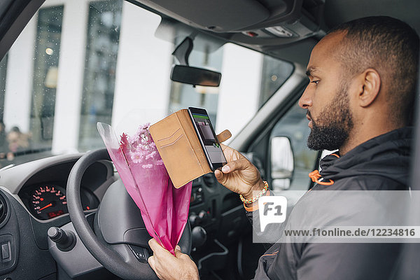 Male messenger with bouquet of flowers looking at smart phone while sitting in delivery van