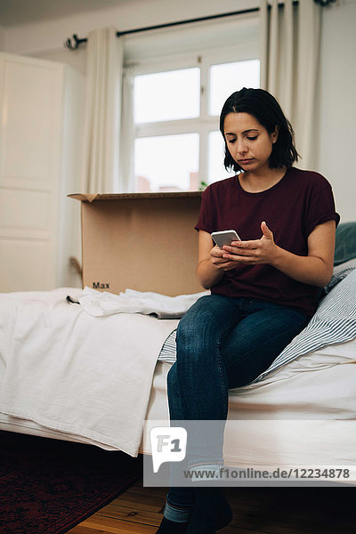Woman using mobile phone while sitting by box on bed