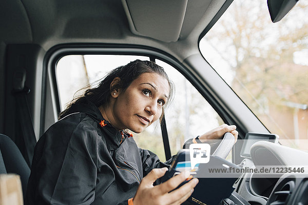 Woman holding mobile phone looking away while driving in delivery van