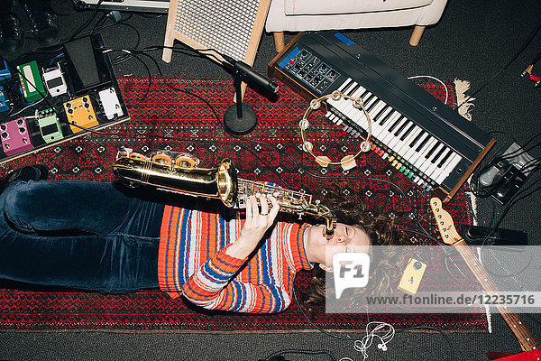 Hochwinkelansicht einer lächelnden Frau die Saxophon spielt während sie im Studio auf dem Teppich liegt.