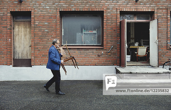 Volle Länge des älteren Mannes  der einen Holzstuhl trägt  während er durch die Werkstatt läuft.