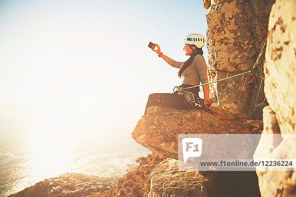 Female rock climber with camera phone photographing sunny ocean view