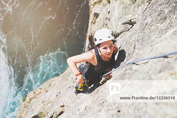 Focused  determined female rock climber scaling rock above sunny ocean