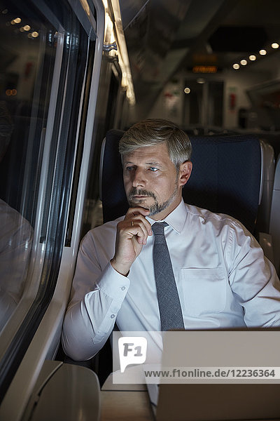 Serious  thoughtful businessman working at laptop  looking out window on passenger train at night