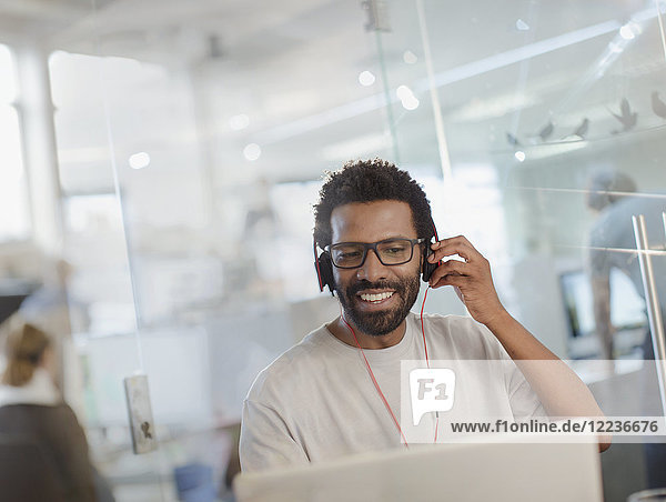 Lächelnder kreativer Geschäftsmann mit Kopfhörern bei der Arbeit am Laptop im Büro