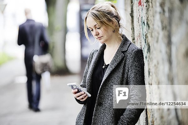 Junge Frau lehnt an einer Wand und schaut auf ihr Smartphone  Köln  Nordrhein-Westfalen  Deutschland  Europa