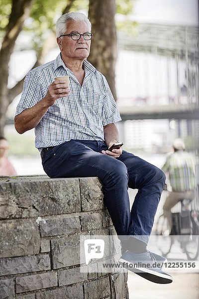 Senior sitzt mit seinem Smartphone und einem Coffee to go auf einer Mauer an der Kölner Rheinuferpromenade  Köln  Nordrhein-Westfalen  Deutschland  Europa