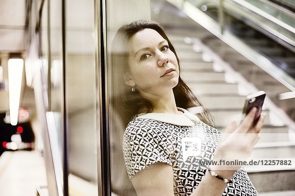 Young woman posing with smartphone behind a glass wall in a subway station  Cologne  North Rhine-Westphalia  Germany  Europe