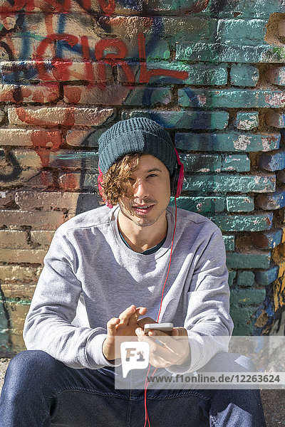 Porträt eines jungen Mannes mit Kopfhörer und Handy vor der Graffiti-Wand