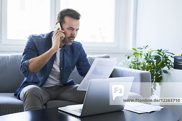 Serious man with documents and laptop on sofa talking on cell phone