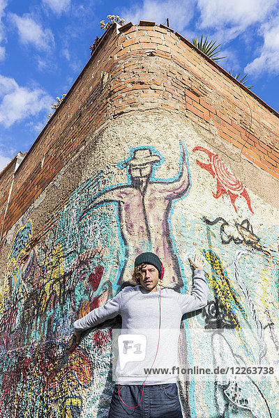 Junger Mann hört Musik mit Kopfhörern  die sich an die Graffiti-Wand lehnen.