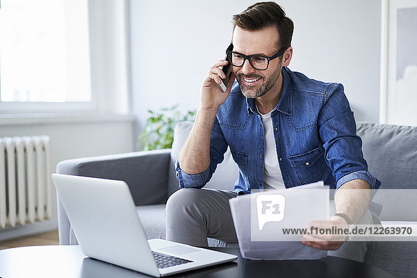 Smiling man with documents and laptop on sofa talking on cell phone