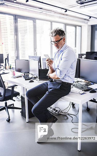 Mature businessman sitting on desk in office using smartphone