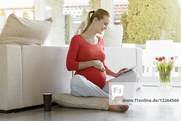 Lächelnde schwangere Frau,  die zu Hause auf dem Boden sitzt und eine Tablette benutzt.