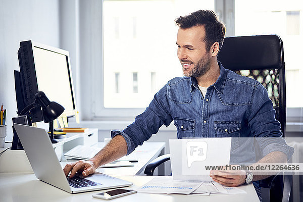 Smiling man holding document and using laptop at desk in office