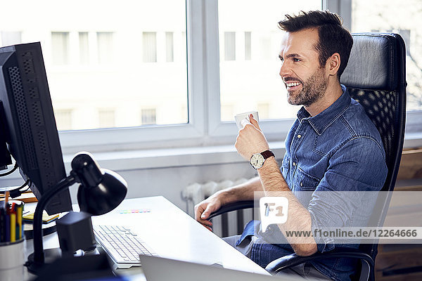Smiling man looking at computer and drinking coffee at desk in office