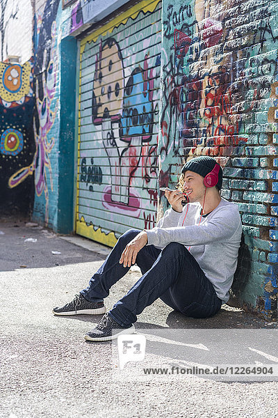 Junger Mann mit Kopfhörer vor der Graffiti-Wand sitzend mit Handy