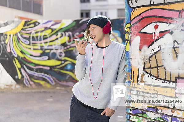 Junger Mann mit Kopfhörer  der sich mit dem Handy an die Graffiti-Wand lehnt.