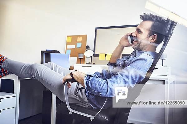 Relaxed man sitting at desk in office talking on cell phone