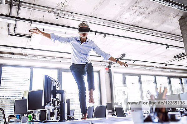 Barefoot mature businessman on desk in office wearing VR glasses
