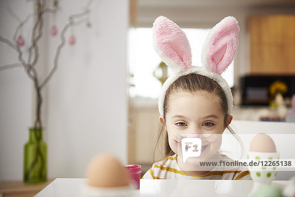 Porträt eines lächelnden Mädchens mit Hasenohren am Tisch mit Ostereiern