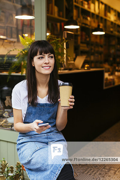 Smiling woman sitting at entrance door of a store holding cell phone and takeaway coffee