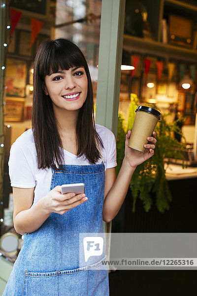 Porträt einer lächelnden Frau mit Handy und Kaffee zum Mitnehmen in der Eingangstür eines Ladens