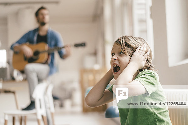 Horrified son covering his ears with father playing guitar at home