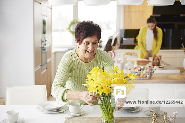 Seniorin arrangiert Blumen auf dem Esstisch mit Familie im Hintergrund