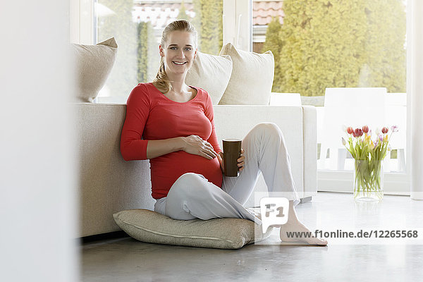 Porträt einer lächelnden schwangeren Frau  die zu Hause auf dem Boden sitzt und eine Tasse hält.