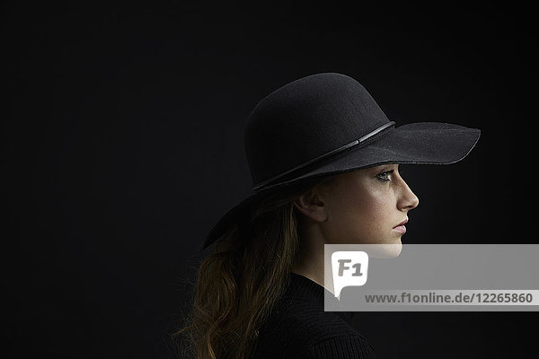 Profil der traurigen jungen Frau mit schwarzem Hut vor schwarzem Hintergrund
