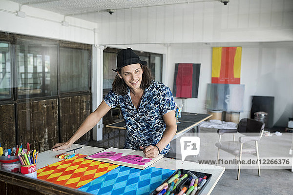 Lächelnder Künstler bei der Arbeit  Zeichnen in einem Notizbuch in seinem Loft-Atelier mit Bildern und Kunstwerken im Hintergrund.