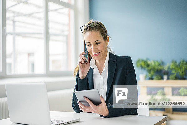 Businesswoman sitting at desk  talking on the phone  looking at digital tablet