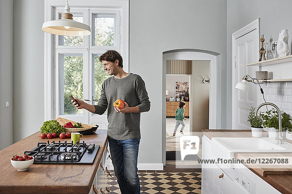 Smiling man using smartphone and holding bell pepper in kitchen