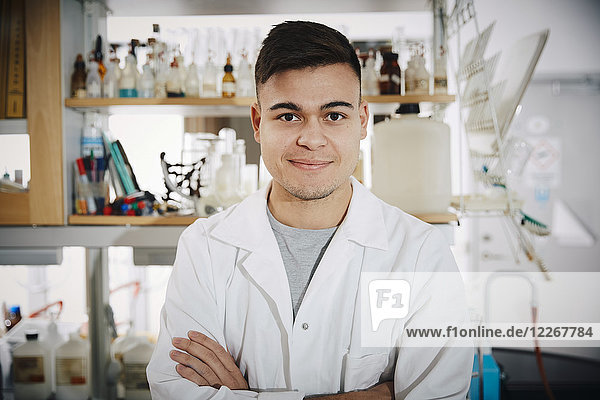 Porträt eines lächelnden jungen Chemie-Studenten  der mit gekreuzten Armen im College-Labor steht.