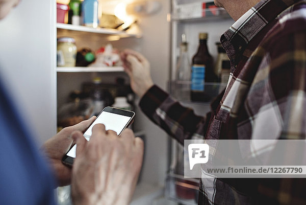 Seniorenfrau mit Smartphone steht in der Küche zu Hause neben dem pensionierten Mann