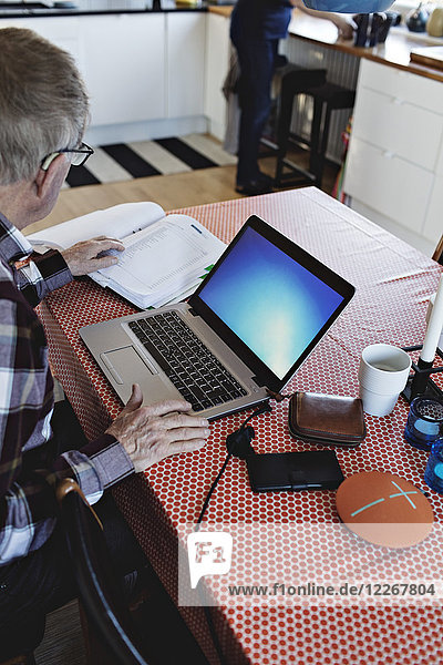 Hohe Winkelansicht des pensionierten älteren Mannes unter Verwendung des Laptops beim Sitzen mit Finanzrechnungen am Esstisch
