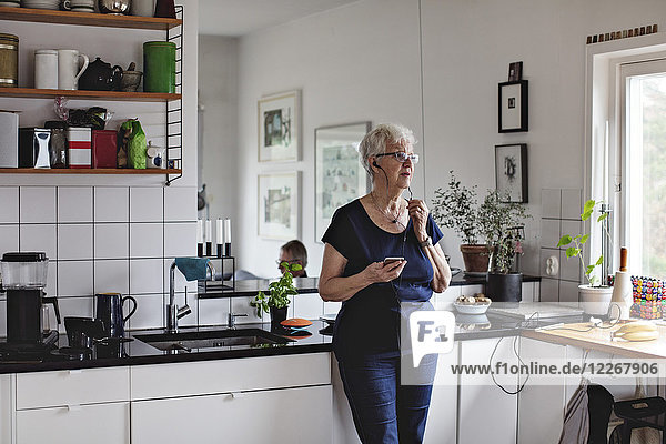 Seniorin im Ruhestand mit Smartphone beim Hören von In-Ear-Kopfhörern in der heimischen Küche