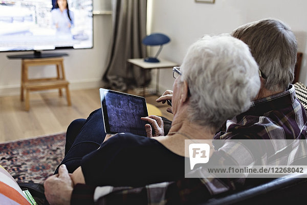 Rear view of senior couple sitting with digital tablet watching TV in living room at home