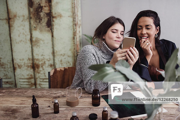 Smiling woman sharing smart phone with female colleague while sitting at table in perfume workshop