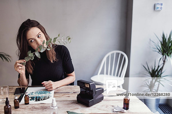 Woman smelling leaves while sitting at table against wall in perfume workshop
