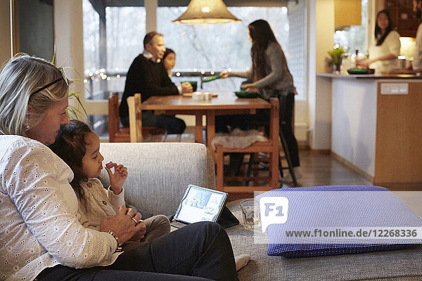 Mädchen und Großmutter beobachten digitales Tablett auf dem Sofa mit Familie im Hintergrund